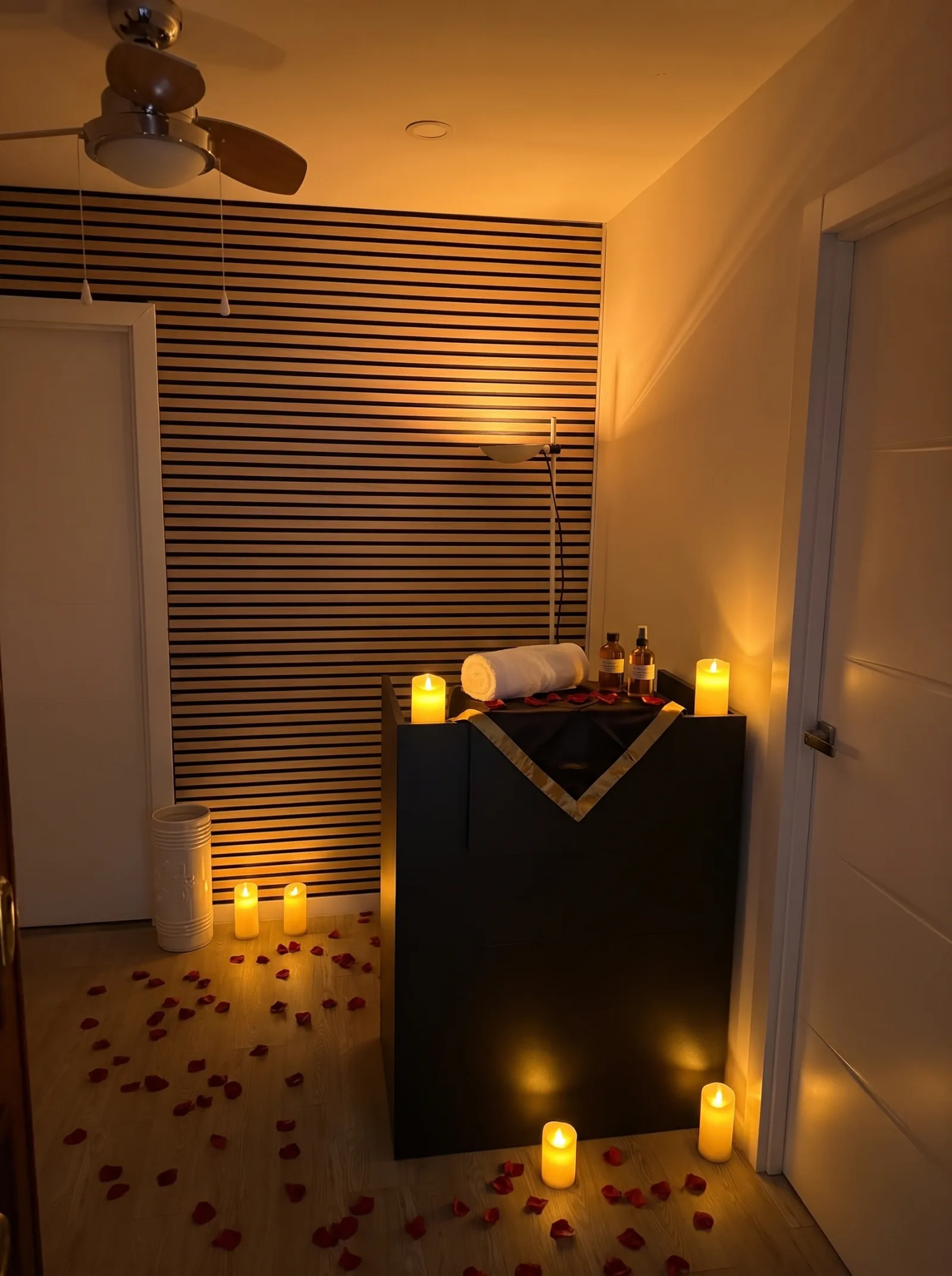 Sala Zen con pared de listones de madera, ventilador de techo, velas encendidas, toalla y frascos de aceite.